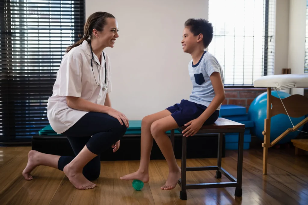 Smiling female therapist kneeling by boy stepping on stress ball at hospital ward